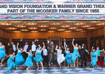 Warner Grand Theatre Exterior Marquee Reads Grand Vision Foundation and Warner Grand Theatre Part of the Mcosker Family Since 1955 While People Jump in Celebration