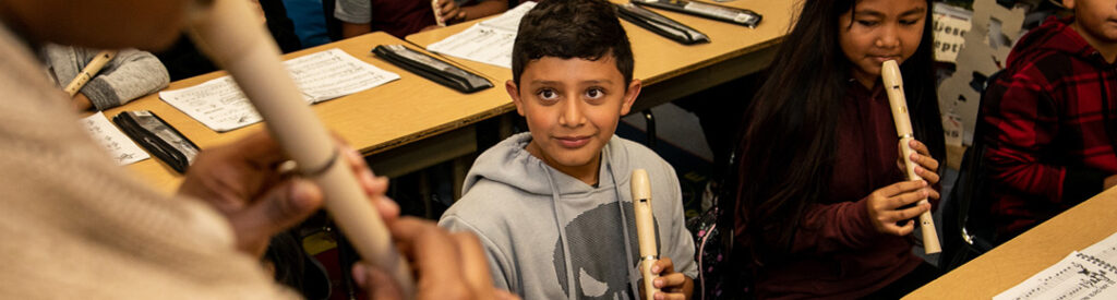 Students at desks play the recorder while a teacher stands in the foreground