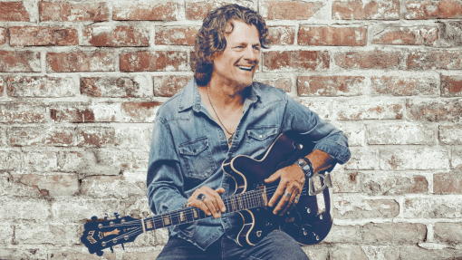 Shawn Jones holding a guitar sitting in front of a brick wall